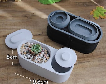May include: Two oval-shaped planters, one white and one black, with gray interior components. The white planter, measuring 19.8 cm long and 8 cm high, contains a succulent and decorative stones. The black planter has two gray inserts. Lids are included.