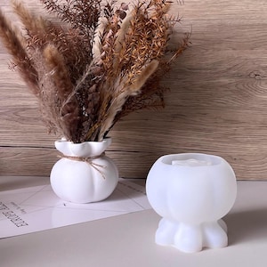 May include: A white ceramic vase in a pumpkin shape, filled with dried brown and beige grasses. Beside it is a white, translucent, pumpkin-shaped candle holder with a three-footed base. The backdrop is a wooden wall.