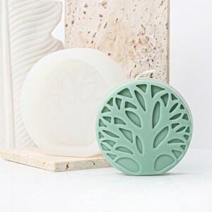 May include: A green candle with a tree design. The candle is round and has a white wick. The candle is sitting on a white surface.