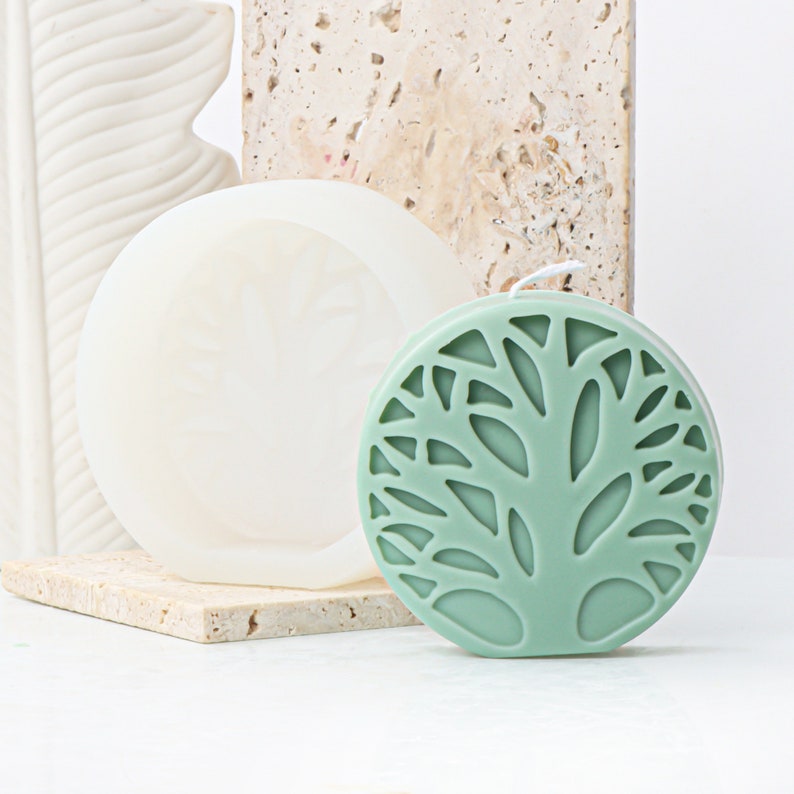 May include: A green candle with a tree design. The candle is round and has a white wick. The candle is sitting on a white surface.