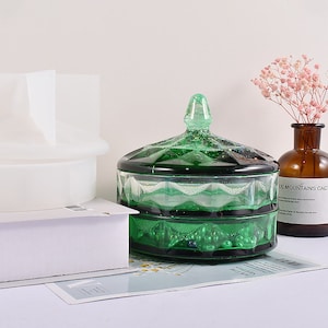 以下が含まれることがあります： 蓋付きの緑色のガラス製の装飾用ジャー。ジャーはダイヤモンド模様で、小さな緑色のビーズで満たされています。ジャーは白い表面の上に置かれています。