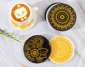 Op de afbeelding: Vier ronde onderzetters met ingewikkelde mandala-ontwerpen. Twee onderzetters zijn zwart met gouden ontwerpen, en twee zijn wit met gouden ontwerpen.