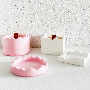 Könnte beinhalten: Zwei Beton-Kerzenhalter mit einem welligen Randdesign. Einer ist rosa und der andere weiß. Beide haben einen Holzdocht.