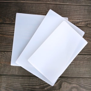 May include: Three white chopping boards of different sizes stacked on top of each other on a brown wooden surface.