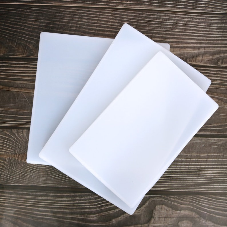 May include: Three white chopping boards of different sizes stacked on top of each other on a brown wooden surface.