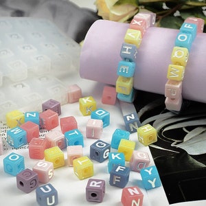 Puede incluir: Surtido de cuentas de letras del alfabeto de colores en varios colores pastel, incluyendo rosa, azul, amarillo y morado. Las cuentas son de forma cúbica y tienen letras blancas en cada lado. Algunas cuentas están enhebradas para formar un brazalete.