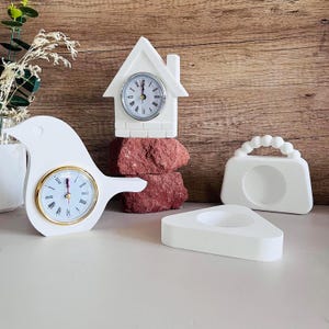 Può includere: Tre figurine di orologio in ceramica bianca. Una è a forma di casa, un'altra a forma di uccello e la terza a forma di borsa. Gli orologi a forma di casa e di uccello hanno lancette e numeri dorati.