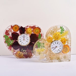 May include: Two heart-shaped resin clocks with dried flowers embedded inside. The clock on the left has red and yellow flowers and the text "Enjoy the Moment". The clock on the right has white and yellow flowers.