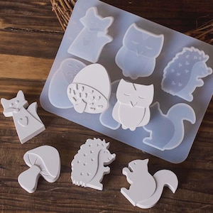 May include: A translucent silicone mold sheet featuring cavities shaped like a fox, owl, hedgehog, squirrel, acorn, and mushroom. Several white, solid, flat-backed resin figures of these same woodland shapes are arranged around the mold.