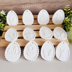 May include: Twelve white ceramic Easter egg-shaped ornaments with various designs, including bunnies, flowers, and a bow. The ornaments have a hole at the top for hanging.