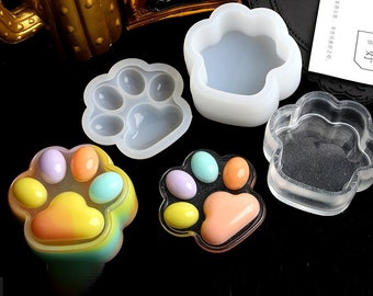 Può includere: Quattro stampi in silicone a forma di impronta di zampa, due sono trasparenti e due sono bianchi. Uno stampo è riempito con resina colorata, creando un'impronta di zampa con cinque punti colorati.