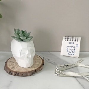 Peut inclure: Un pot de fleurs en forme de crâne géométrique blanc avec une plante succulente verte à l'intérieur, posé sur une tranche de bois. Un petit calendrier avec un tigre de dessin animé et le texte "2022" est en arrière-plan.