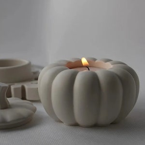 May include: A white ceramic pumpkin-shaped candle holder with a lit candle inside. The candle holder has a ribbed design and is sitting on a white surface. Other ceramic pieces are in the background.