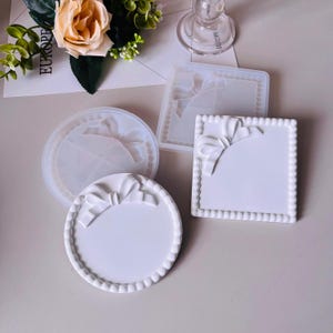 Peut inclure: Un ensemble de moules en résine blanche et de dessous de verre finis. Les moules sont de forme carrée et ronde, avec un motif de nœud. Les dessous de verre finis sont blancs avec un bord perlé et un détail de nœud. Un arrangement floral et le mot "EUROPE" sont visibles.