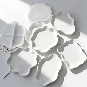 May include: A collection of white ceramic plates with a unique, organic, wavy edge design. The plates vary in shape and size, with some featuring divided sections. The plates are arranged on a white surface, with natural light casting shadows.