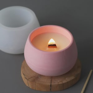 May include: A lit pink candle in a round ceramic container with a wooden wick, on a small wooden slab. The candle's wax is creamy, with another white container in the background.