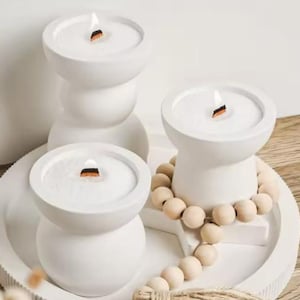 Peut inclure: Trois bougeoirs en céramique blanche avec une mèche en bois dans chacun. Les bougeoirs sont sur un plateau avec une guirlande de perles en bois.
