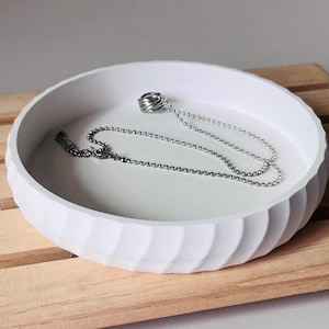 Op de afbeelding: Een witte keramische schaal met een golvend ontwerp, met een zilveren ketting met een zilveren ringhanger.