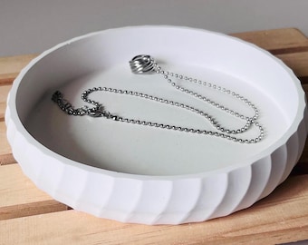 May include: A white ceramic dish with a wavy design, featuring a silver chain necklace with a silver ring pendant.