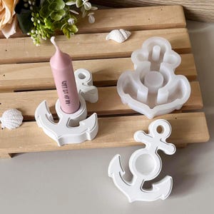 Könnte beinhalten: Weiße Anker-Kerzenhalter und eine Silikonform auf einer Holzoberfläche. Eine rosa Kerze mit Text befindet sich in einem Halter. Dekorative Muscheln sind ebenfalls vorhanden. Nautisch thematisierte Dekoration.