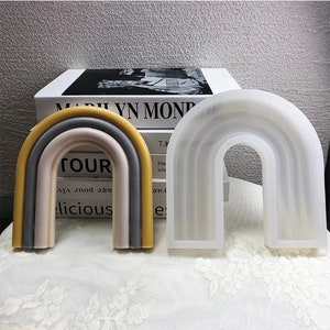 May include: Two rainbow-shaped silicone molds. The mold on the left is multi-colored with yellow, gray, and pink. The mold on the right is white. Both molds are shaped like a rainbow arch.