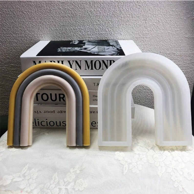 May include: Two rainbow-shaped silicone molds. The mold on the left is multi-colored with yellow, gray, and pink. The mold on the right is white. Both molds are shaped like a rainbow arch.