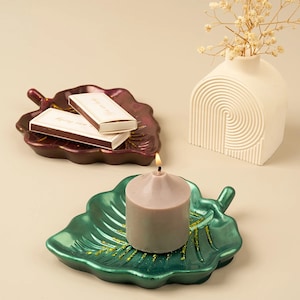 Pode incluir: Dois porta-velas de cerâmica em forma de folha, um bordô e outro verde, com uma vela cinza acesa no porta-velas verde. O porta-velas bordô tem duas caixas de fósforos no topo.