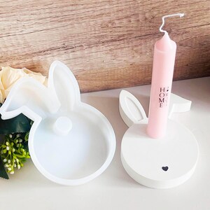 Może przedstawiać: Biała silikonowa forma w kształcie królika z małym sercem na dole. Forma jest otwarta i pokazuje wnętrze formy. Obok formy znajduje się biały ceramiczny świecznik w kształcie królika z różową świecą w środku. Świecznik ma napis "HOME".