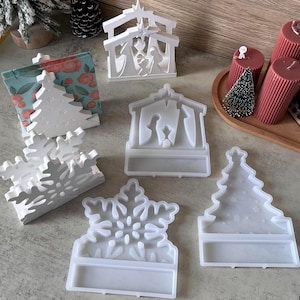 May include: A collection of white Christmas-themed decorations, including a snowflake, Christmas tree, and Nativity scene. The decorations are made of a solid material and are designed to stand upright. The image also includes a small Christmas tree and candles.