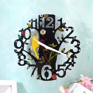 Könnte beinhalten: Schwarze Uhr in Form eines Baumes mit einem gelben Vogel und echten gepressten Blumen. Das Zifferblatt der Uhr hat römische Ziffern und einen weißen Zeiger.