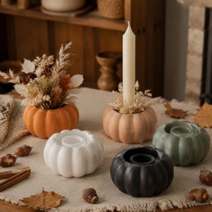 May include: A collection of pumpkin-shaped candle holders in various colors: orange, peach, white, black, and sage green. Some holders contain dried flowers or a white taper candle. Acorns and cinnamon sticks are scattered around the holders on a neutral-colored tablecloth.