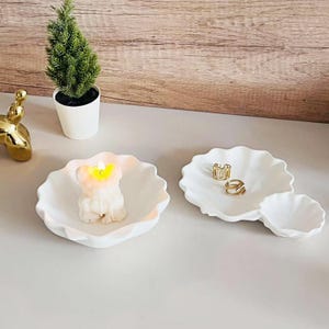 Op de afbeelding: Twee witte keramische schalen met gegolfde randen. Een schaal heeft een kleine witte kaars in de vorm van een beer. De andere schaal heeft twee gouden ringen.