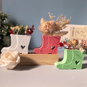May include: Three colorful boot-shaped planters, each filled with dried flowers. The planters are white, pink, and green, with a heart-shaped cutout and dotted pattern. They sit on a wooden block.