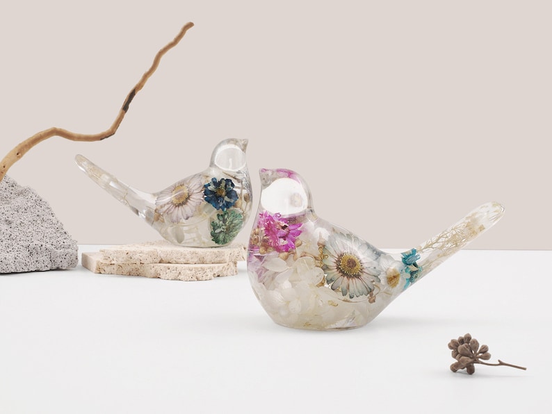 May include: Two clear glass bird figurines with dried flowers and leaves inside. The birds are facing each other and are on a white surface.
