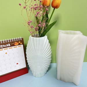 Peut inclure: Deux vases en céramique blanche avec un motif texturé en forme d'éventail. Le vase de gauche est de forme ovale et contient un bouquet de gypsophile rose et de tulipes orange. Le vase de droite est plus haut et rectangulaire. Un calendrier brun avec le texte "正在认真生活" est en arrière-plan.