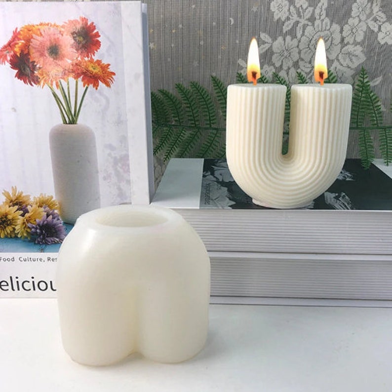 May include: Two white pillar candles with a unique U-shaped design. The candles are lit and have a warm glow. The candles are on a white surface with a book and a vase of flowers in the background.