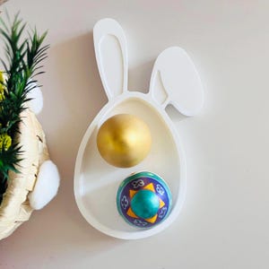 May include: White ceramic bunny-shaped dish with two Easter eggs. One egg is gold and the other is green with purple and yellow designs.