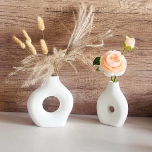 May include: Two white ceramic vases with unique, modern designs. The vase on the left has a large, circular opening, while the vase on the right has a smaller, oval opening. The vase on the left is filled with dried pampas grass, while the vase on the right is filled with a single peach-colored rose.
