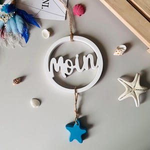 Op de afbeelding: Een witte houten cirkel met het woord "moin" in cursief geschreven. Een blauwe ster hangt aan de onderkant van de cirkel.