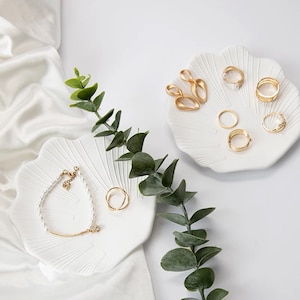 Può includere: Due piatti in ceramica bianca a forma di conchiglia, uno con un braccialetto di perle e un anello d'oro, l'altro con un paio di orecchini d'oro e diversi anelli d'oro.