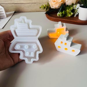 Könnte beinhalten: Eine transparente, flexible Silikonform in Schiffsform, gehalten von einer Hand. Zwei weiße schiffsförmige Kerzenhalter mit leuchtenden orangefarbenen Lichtern befinden sich neben der Form. Die Kerzenhalter haben kreisförmige Aussparungen.