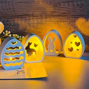 Puede incluir: Cuatro portavelas de madera blanca en forma de huevo de Pascua con diferentes recortes. Los portavelas están iluminados con luces LED blanco cálido. Los recortes incluyen un conejito, una mariposa y corazones.