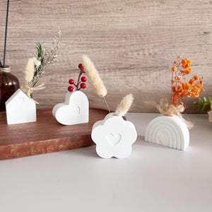 May include: Four white ceramic vases in different shapes, including a house, heart, flower, and rainbow, with dried flowers in each.