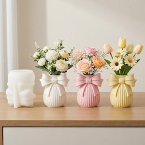 May include: A collection of four decorative vases. Three vases are pastel colors: white, pink, and yellow, each with a bow design. The vases hold bouquets of flowers. The fourth vase is white and has a unique sculpted design.