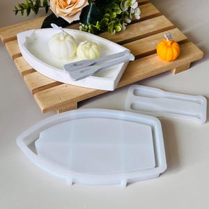 May include: A white boat-shaped tray with two small white pumpkins and grey oars. An orange pumpkin candle sits on a wooden pallet. Boat and oar-shaped silicone moulds are also present.