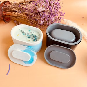 May include: Two oval-shaped silicone molds for making candles. One mold is white with blue swirls and has a dried flower arrangement inside. The other mold is black with a gray silicone insert.