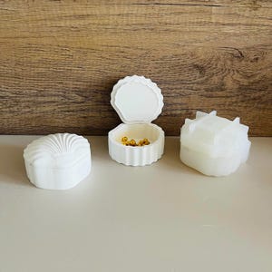 May include: Three white ceramic trinket boxes. One box is shaped like a seashell, another is a round box with a lid, and the third is a geometric box with a lid.