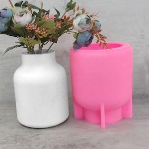 May include: A white vase with a slight taper holds a bouquet of flowers with blue, pink, and green hues. Next to it is a bright pink, cylindrical vase with three small legs, designed for floral arrangements.