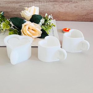 Peut inclure: Trois tasses en céramique blanche en forme de cœur avec des poignées. Les tasses sont disposées sur une surface en bois brun clair.