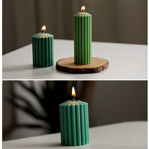 May include: Three green pillar candles with a ribbed design. The candles are lit and have a warm glow. The candles are on a white surface.